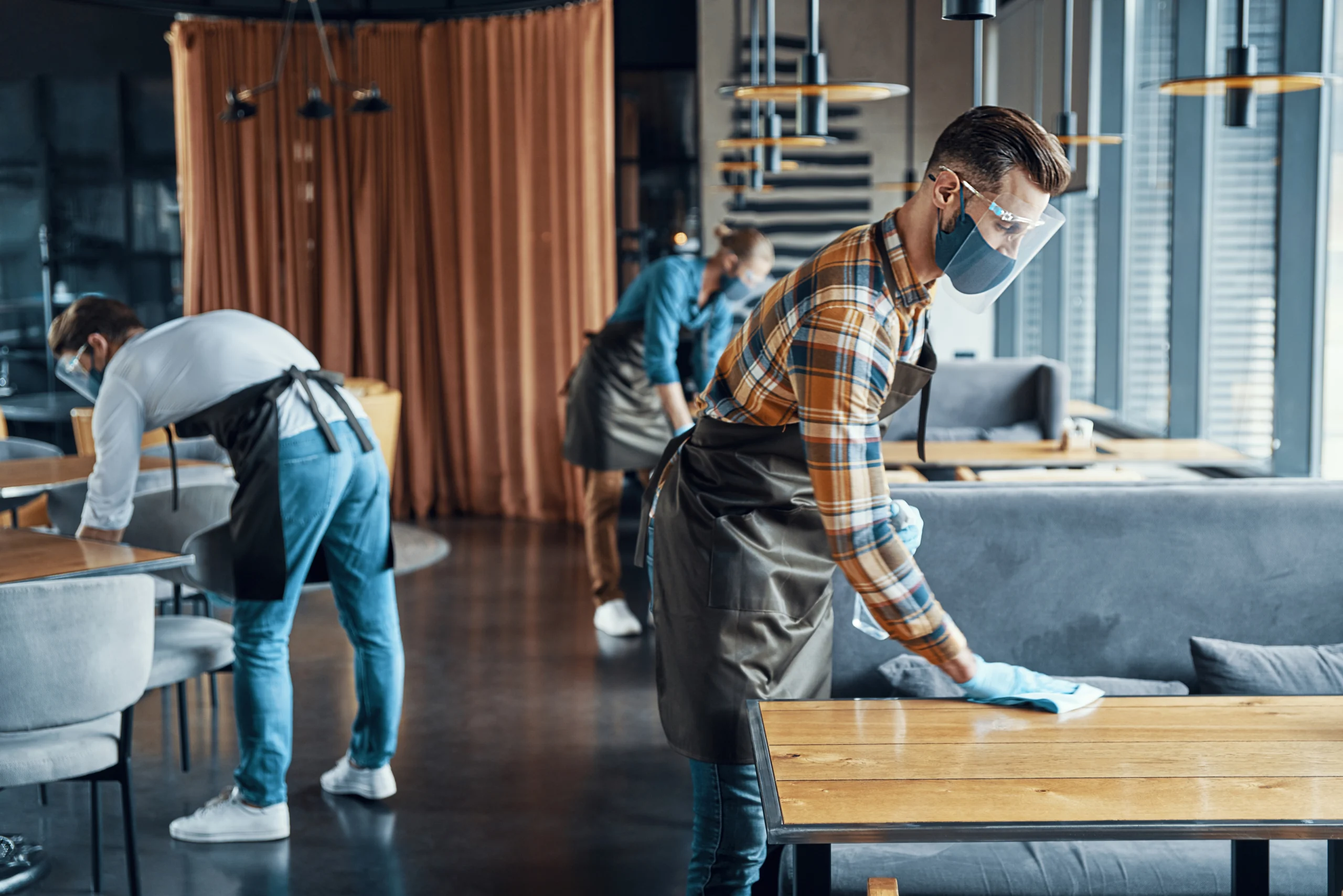 hombre joven responsable camarero en protector facial rociar desinfectante en las mesas en el restaurante convert.io scaled